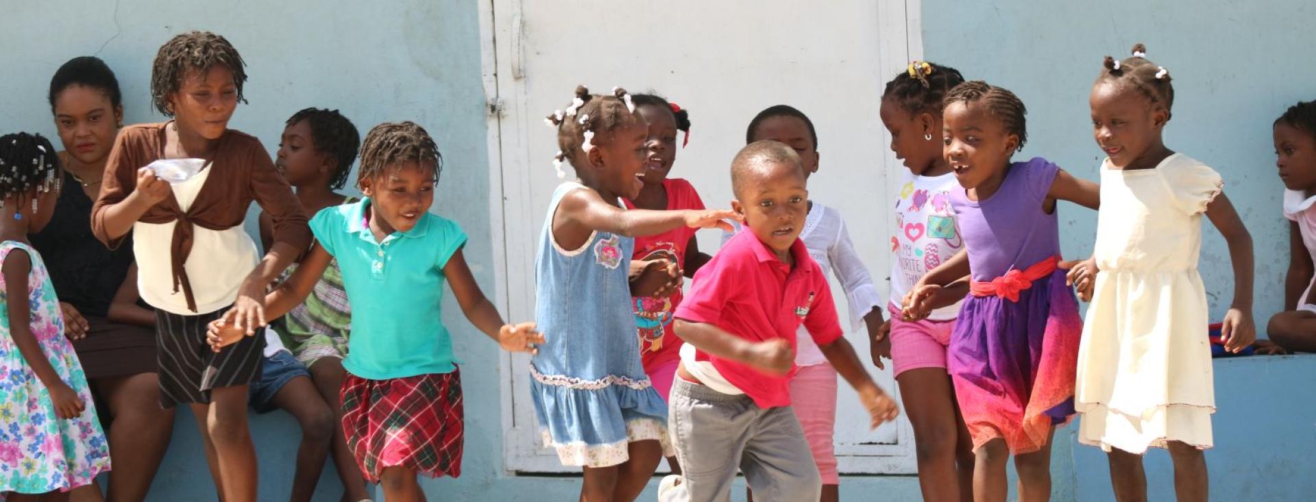 An action shot of several young joyful Black children holding hands in front of a pale blue wall