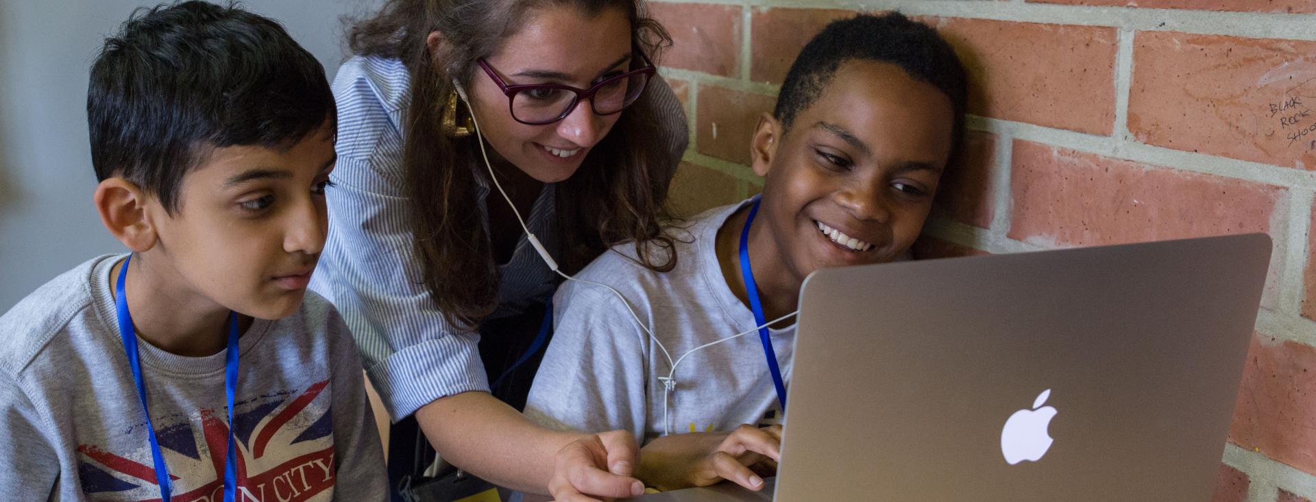 A young woman leans over two boys who are using a Macbook