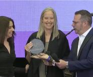 A white woman with long blond hair receives an award from a white man in a suit and a white woman with brown hair 