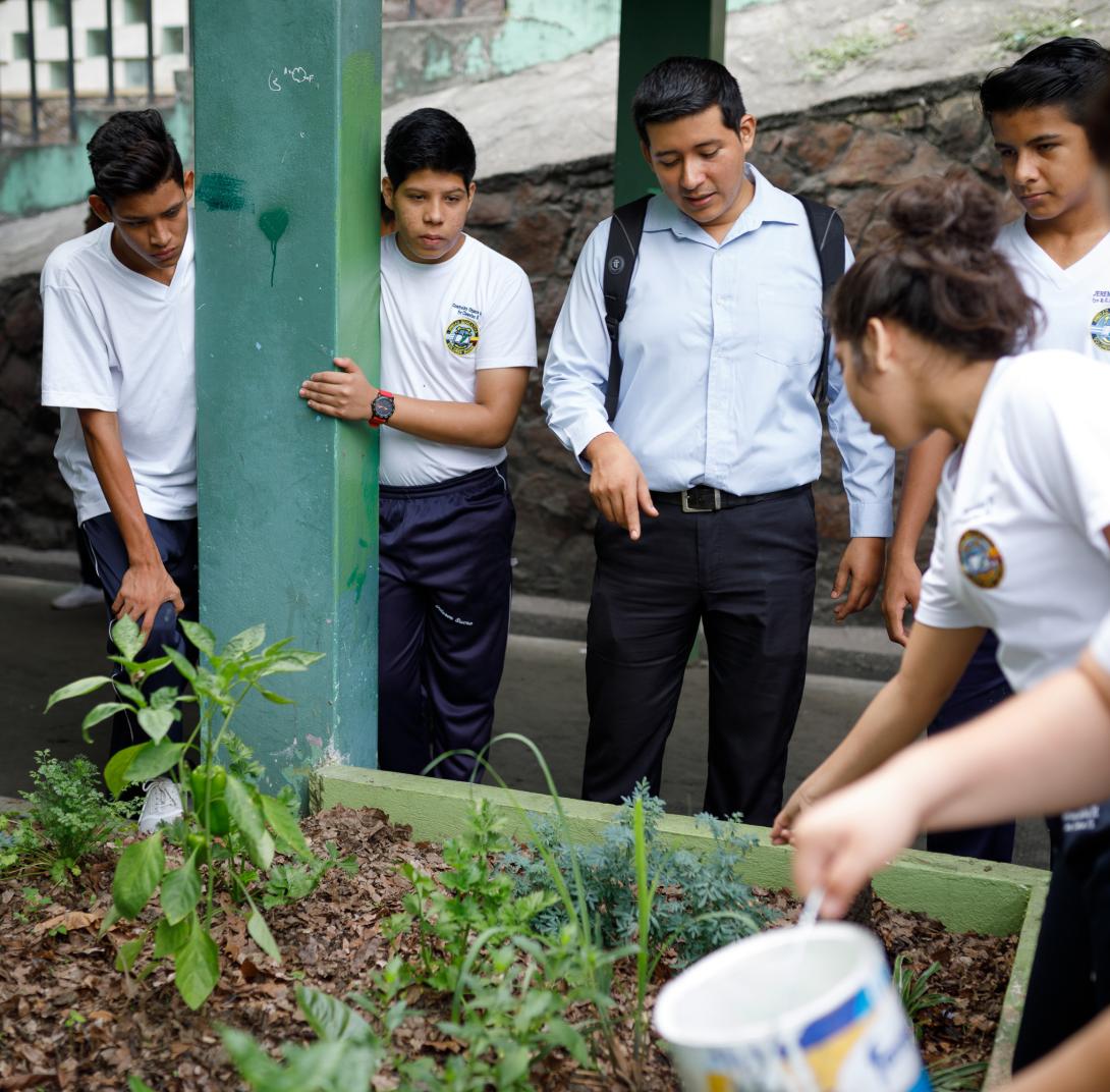 A young male teacher speaks to a small group of teenage students in school uniforms as they tend to a small raised garden bed