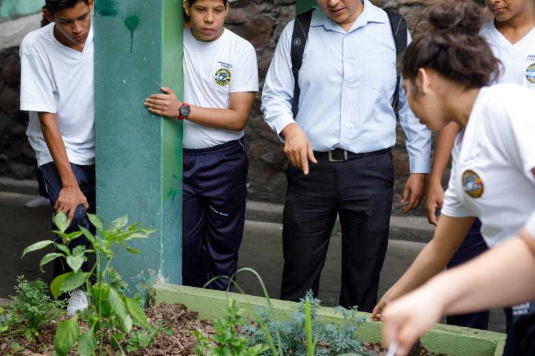 A young male teacher speaks to a small group of teenage students in school uniforms as they tend to a small raised garden bed