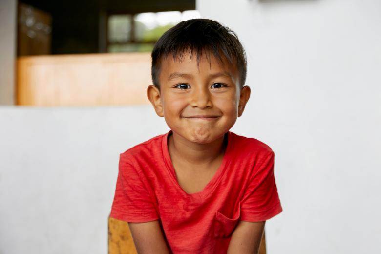 A smiling boy with brown skin and wearing a red t-shirt looks right into the camera
