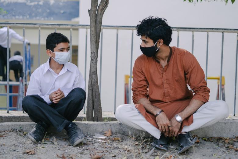A pre-teen boy sits on a curb next to a young man. The two face each other, both wearing masks and sitting cross-legged.