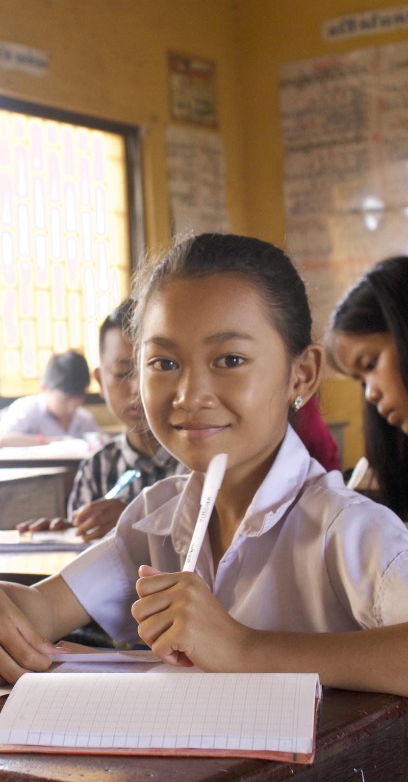 A young female Asian student is seated at a desk in a classroom, smiling as she looks directly into the camera