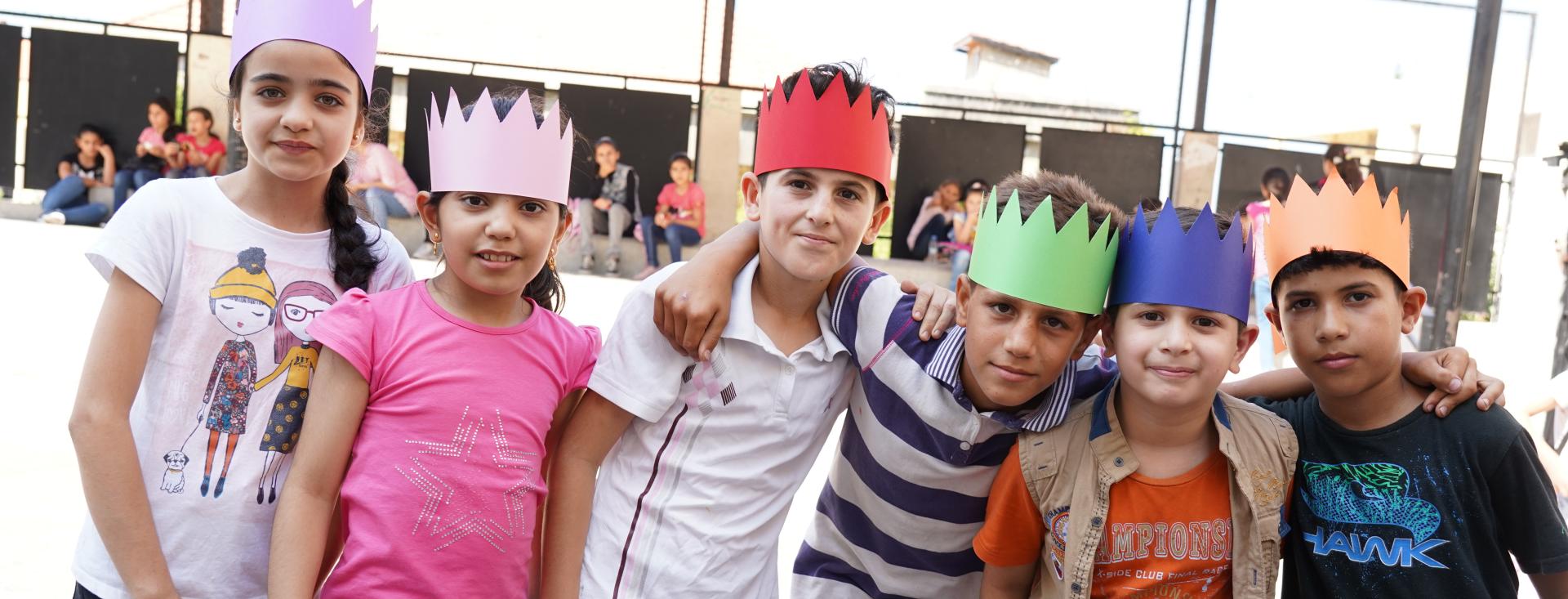 A group of 7 children wearing multi-colored paper crowns stand with their arms around each other's shoulders looking into the camera