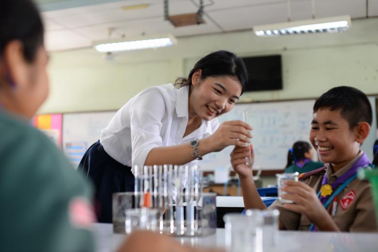 An Asian female teacher smiles as she hands a test tube to an Asian boy in a school uniform who is also smiling. There are more test tubes in a rack on the table