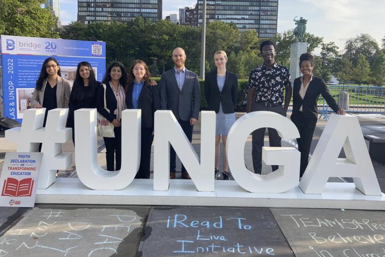 A diverse group of 8 young adults stand behind a three dimensional sign that says UNGA