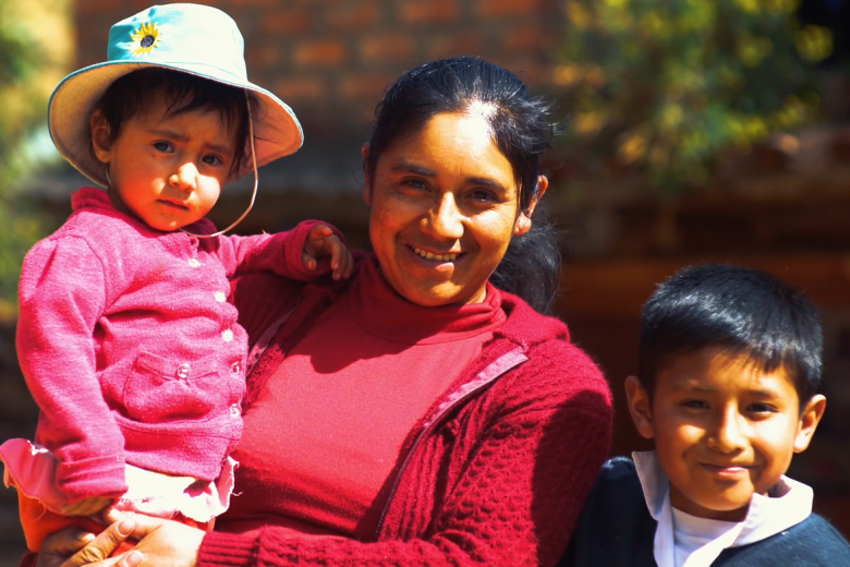 A brown-skinned woman in a red sweater holding a baby wearing a hat smiles at the camera alongside a small boy