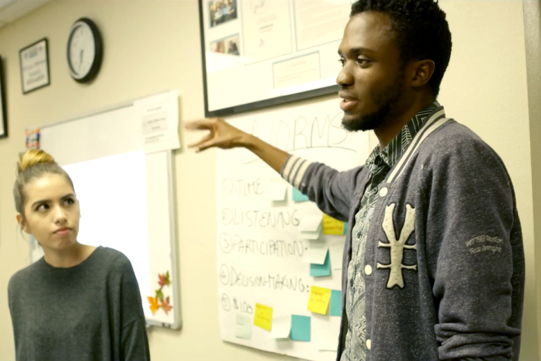 A young Black man speaks with his back against a white board as a young woman with a bun listens